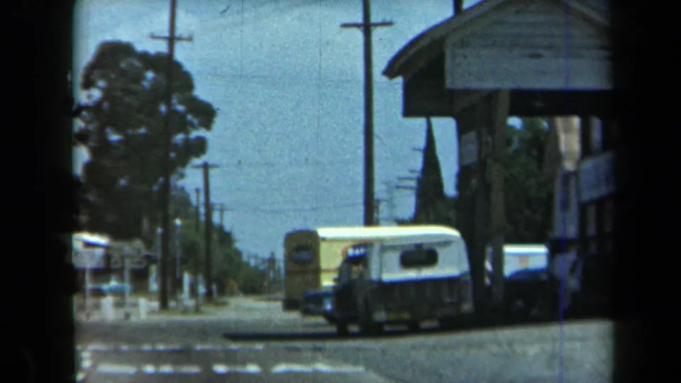 1969 California 8mm frame showing a yellow and white van parked along a quiet road near a storefront, utility poles, and trees.