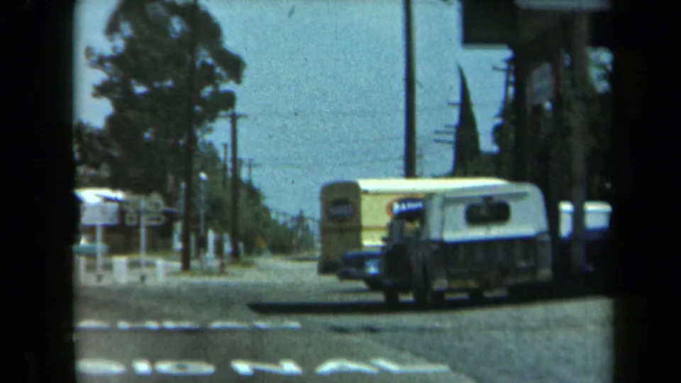 1969 California roadside frame with a parked van, distant sign, utility poles, trees, and the edge of a small storefront.