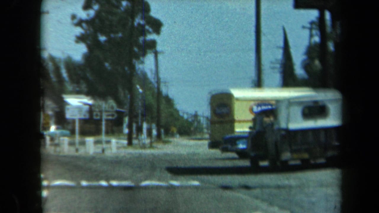 1969 California home-movie view of a road intersection with a yellow step van crossing, highway signs, cars, trees, and utility poles.