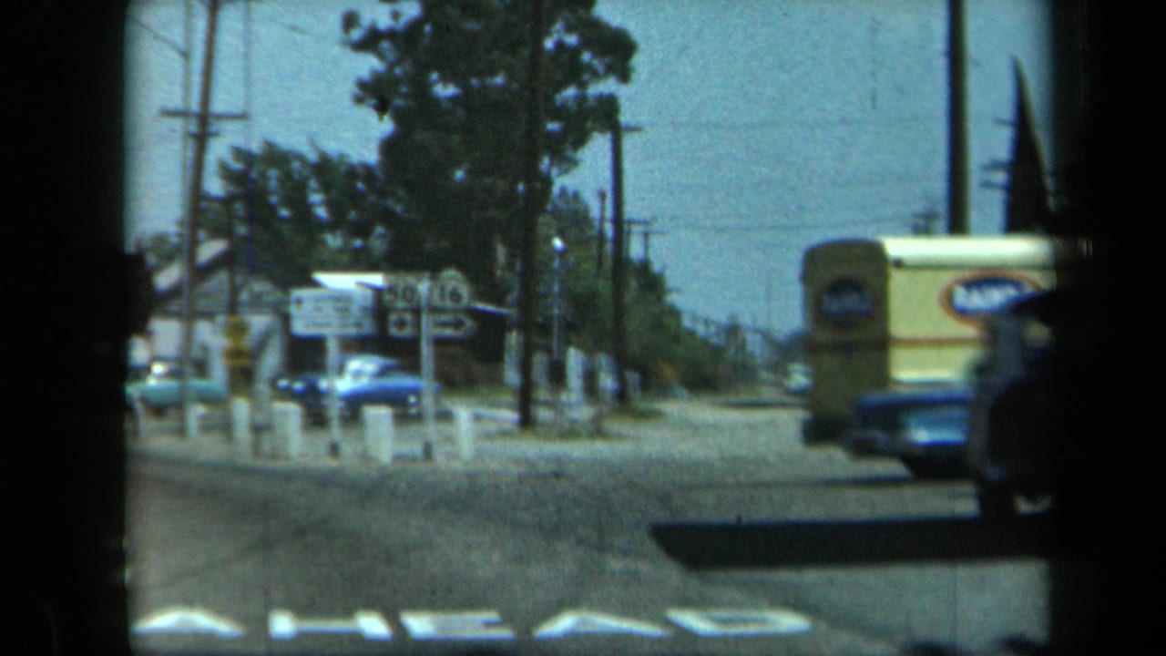 Intersection view with route signs, a blue car, a yellow vehicle edge, utility poles, trees, and low buildings.