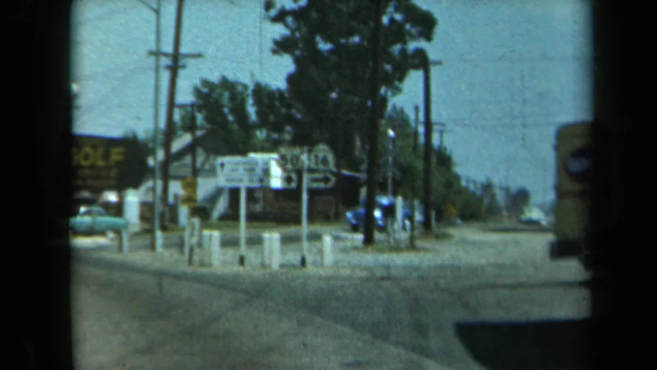 1969 California street scene with a prominent GOLF sign, highway markers, a green car, utility poles, trees, and roadside storefronts.
