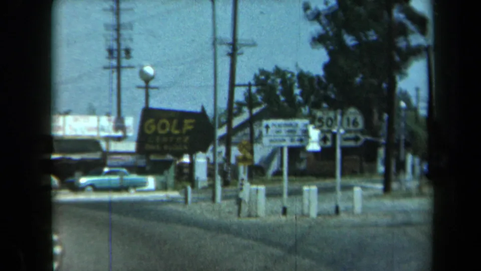 1969 California home-movie frame with a GOLF sign, highway markers, a pale blue car, utility poles, trees, and low roadside buildings.