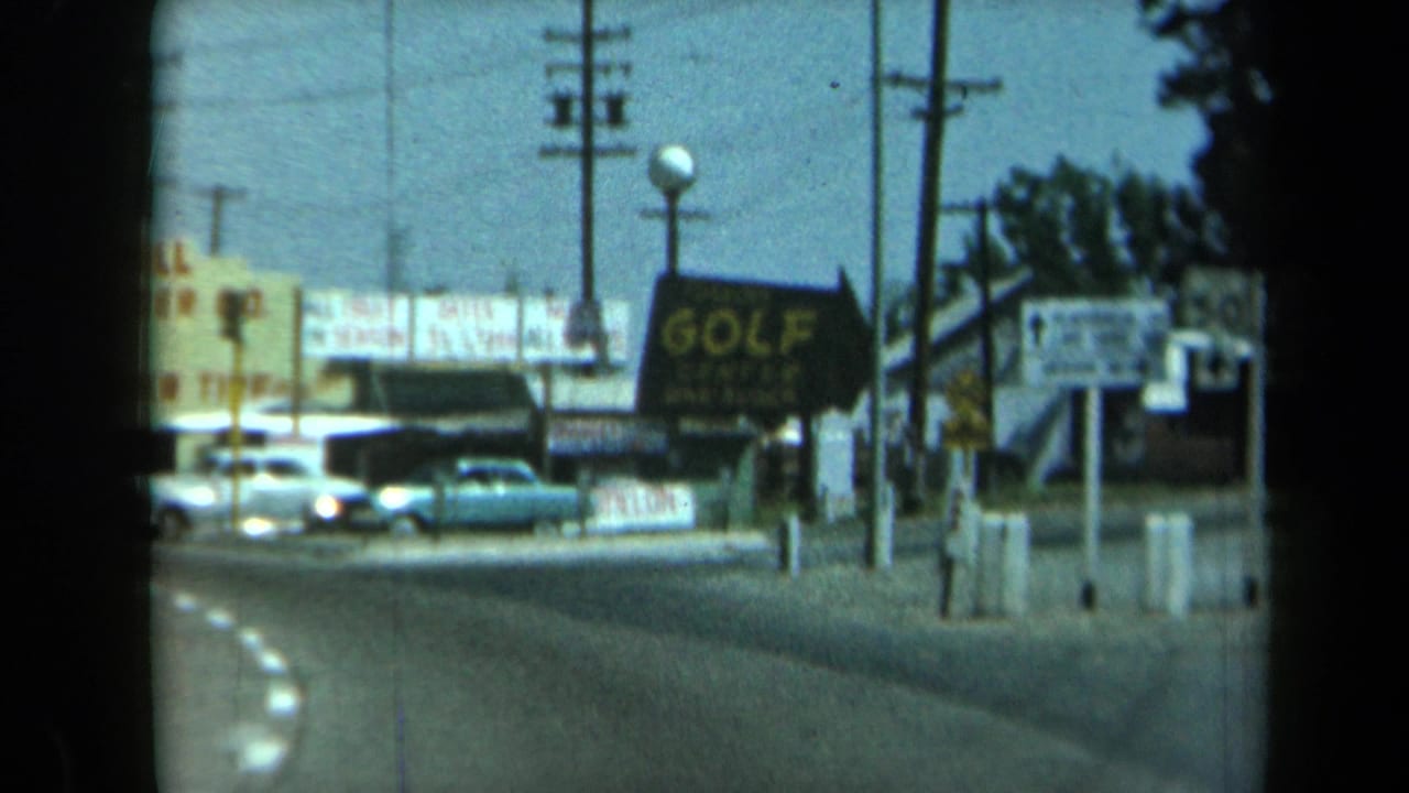 1969 California 8mm frame showing a curved road, parked classic cars, utility poles, a golf sign, and roadside commercial signage.