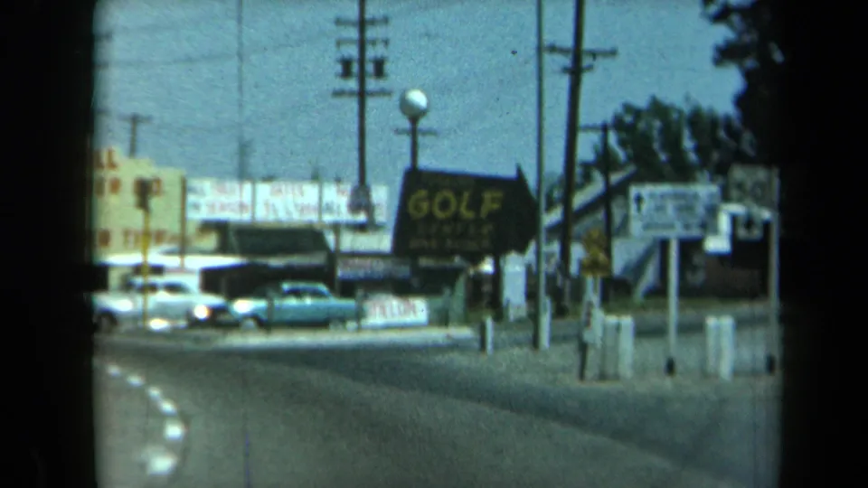1969 California 8mm frame showing a curved road, parked classic cars, utility poles, a golf sign, and roadside commercial signage.