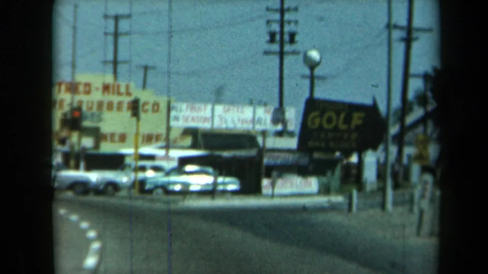 1969 California street corner with parked American cars, a large golf sign, utility poles, and visible roadside business signs.