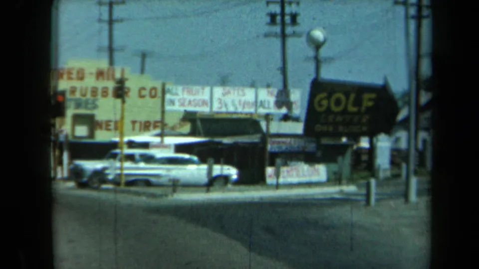 1969 California 8mm frame with classic cars parked along a roadside commercial corner under utility wires and large signs.