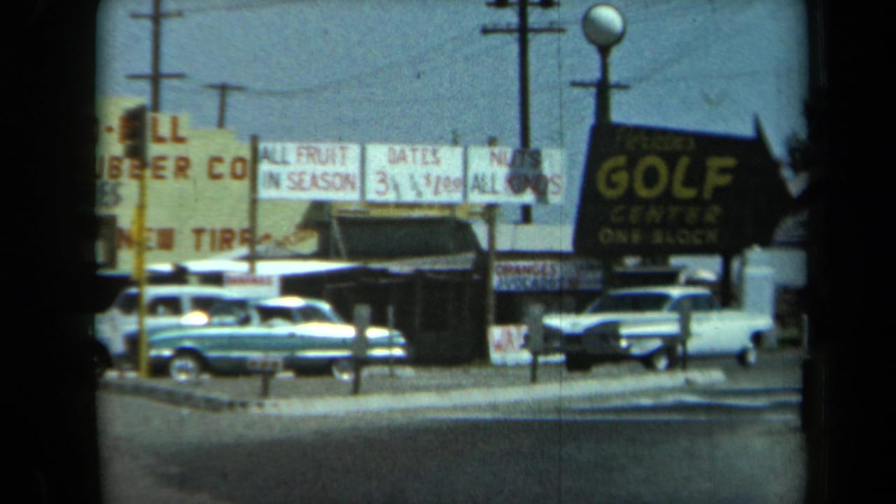 Roadside shops with vintage cars parked near signs for fruit, tires, dates, nuts, and golf.