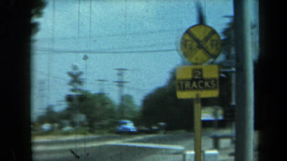 A California road scene from the 1969 home movie shows a railroad crossing sign marked two tracks, with cars, poles, and roadside trees behind it.