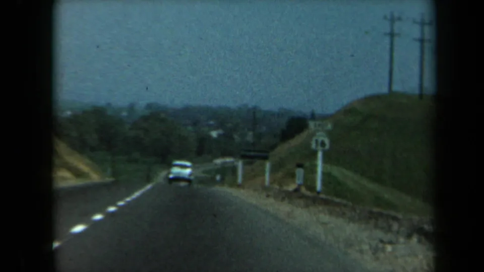 A white car travels on a rural California highway, with roadside signs, utility poles, and low hills visible through the 8mm frame.