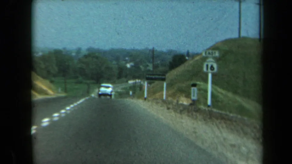 The home-movie view looks down a California highway as a white car passes a route marker, hillside, utility poles, and trees.