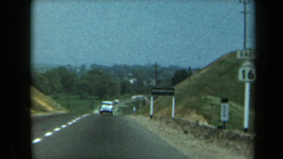 A white car drives along the rural California highway near a hillside, visible route marker, utility poles, and roadside sign.
