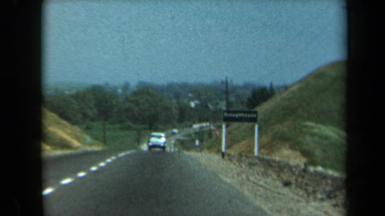 1969 California 8mm view from a moving car on a rural road with hills, utility poles, a vehicle ahead, and a roadside sign.