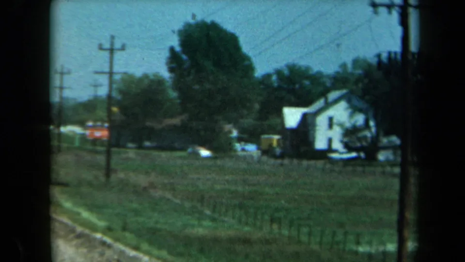 1969 California home-movie frame showing a rural field, roadside utility poles, trees, and a white farmhouse from a moving car.