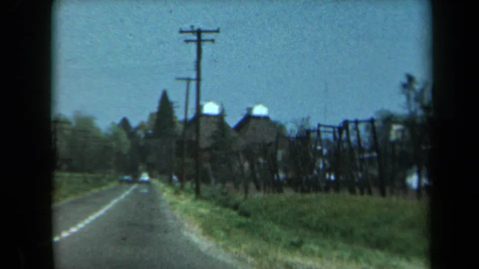 1969 California rural road scene with a car ahead, utility poles, grassy shoulder, and dark wooden structures beside the road.