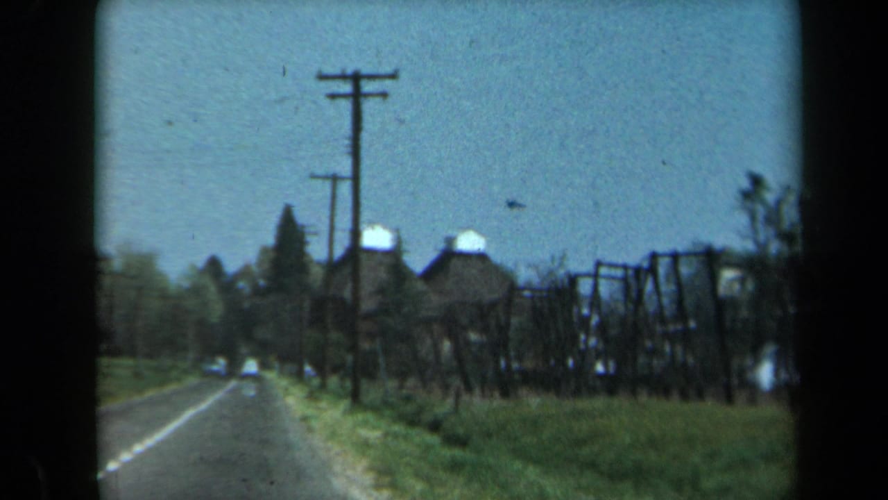 1969 California home-movie frame showing a two-lane road, grassy shoulder, utility poles, and rural buildings in the distance.