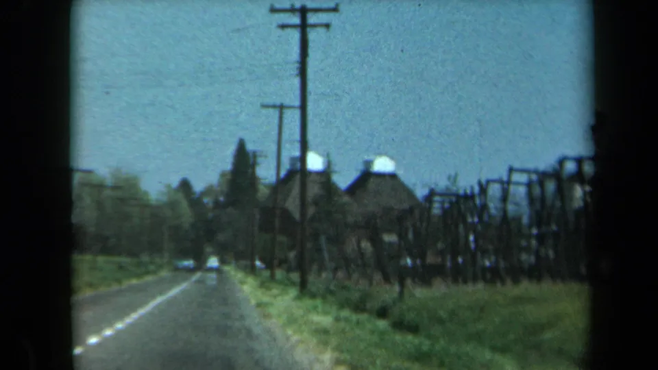 Vehicle-shot 1969 California road view with utility poles, grassy verges, rustic roadside structures, and low buildings ahead.