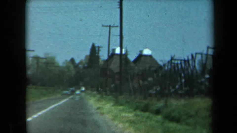 1969 California roadside frame with a two-lane road, utility poles, grassy shoulder, and dark trellis-like structures beside rural buildings.