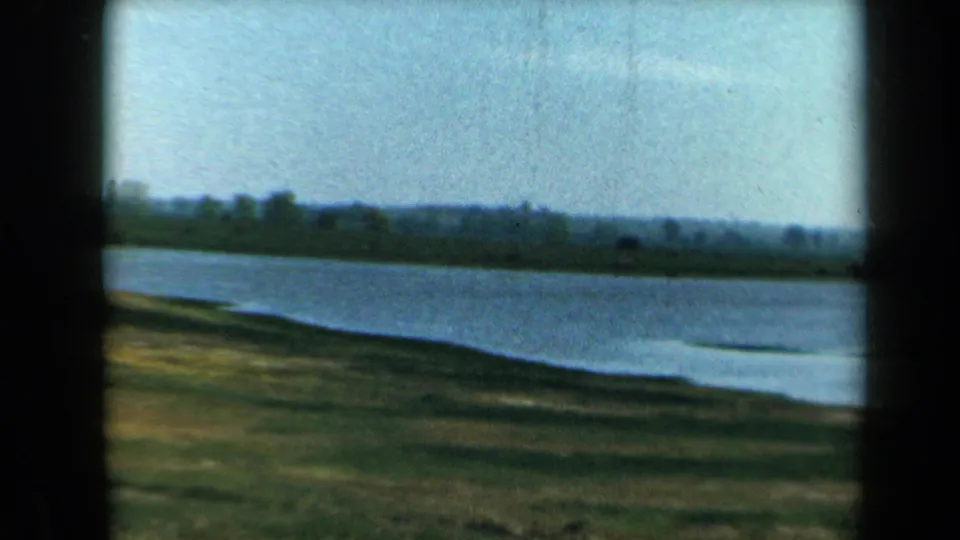 1969 California home-movie frame revealing a calm lakeshore with grassy bank, distant trees, low hills, and bright sky.