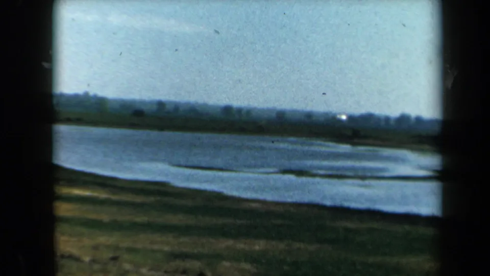 1969 California 8mm frame showing rippled lake water, grassy bank, distant trees, and pale sky with visible film texture.