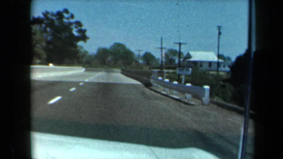 1969 California windshield view down a two-lane road with utility poles, curb markers, trees, and a small white roadside building.