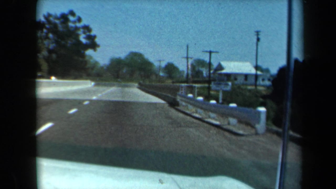 1969 California 8mm view from a vehicle approaching a small bridge with a roadside house, utility poles, trees, and blue sky.