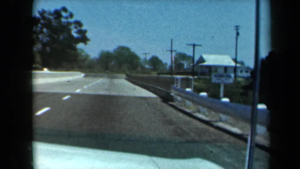 1969 California road view as the vehicle starts across a small bridge, with a white roadside house and utility poles ahead.