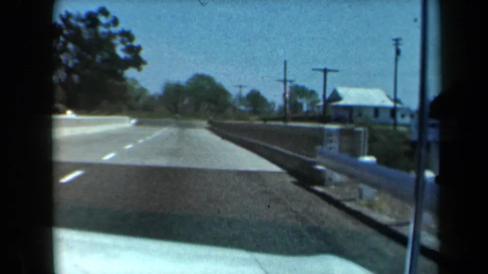 1969 California 8mm frame showing a moving-car view over a bridge toward a quiet road lined with utility poles and trees.