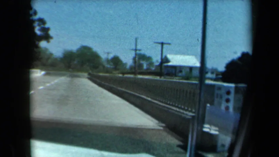 1969 California vehicle POV near the end of a small bridge, showing the guardrail, roadside house, utility poles, and open sky.