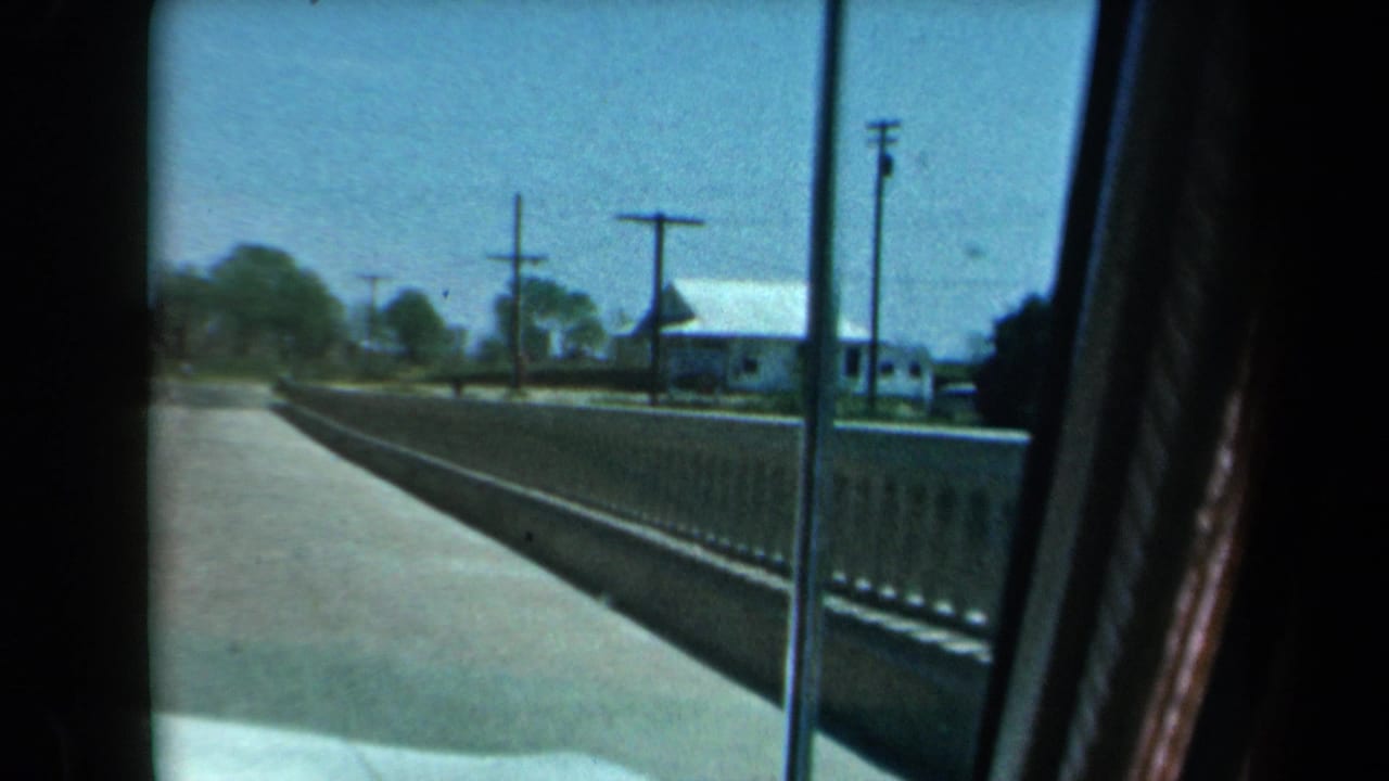 1969 8mm California road-trip view from inside a moving car, showing a rural roadside, railing, utility poles, and distant buildings.
