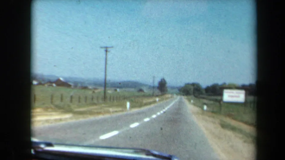1969 8mm California driving view from a car windshield, showing a two-lane road through open fields with hills in the distance.