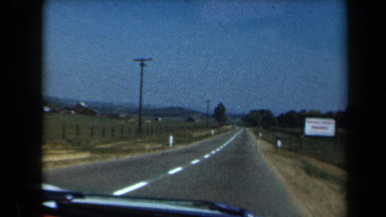 Rural two-lane road seen through a car windshield, with fields, fences, utility poles, and distant hills.