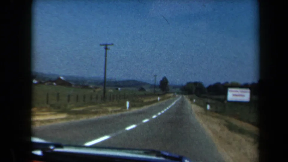 1969 California 8mm frame showing a forward car view along a quiet rural road bordered by fields, fencing, utility poles, and low hills.