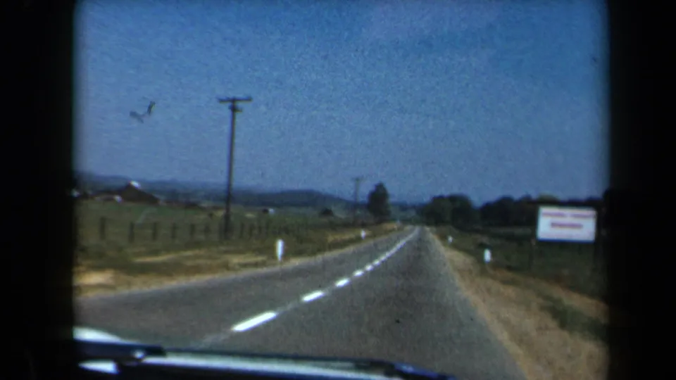 1969 California 8mm frame of a car-view road scene with dashed lane markings, dry shoulders, utility poles, fields, and a roadside sign.