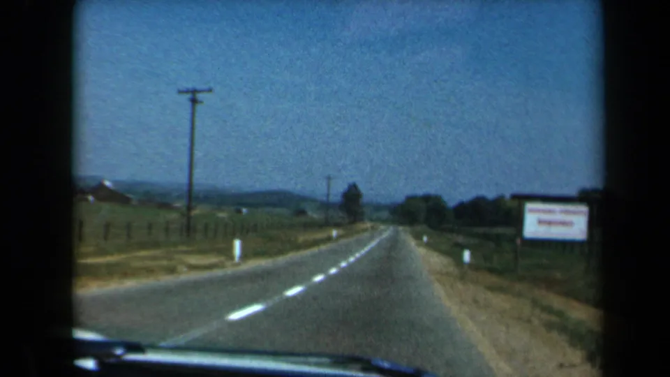 1969 California 8mm frame showing the road ahead from inside a moving car, with rural fencing, a roadside sign, fields, and low hills.