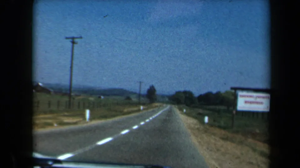 1969 California 8mm frame with a forward driving view of a country road curving past fields, fences, utility poles, and a sign.