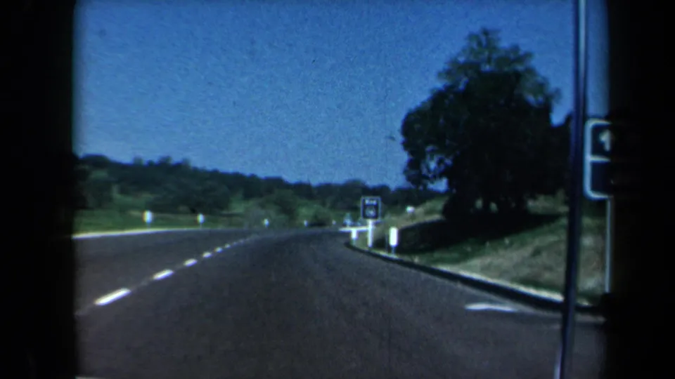 In-car 8mm view of a California highway curve with roadside signs, trees, hills, and a clear blue sky.