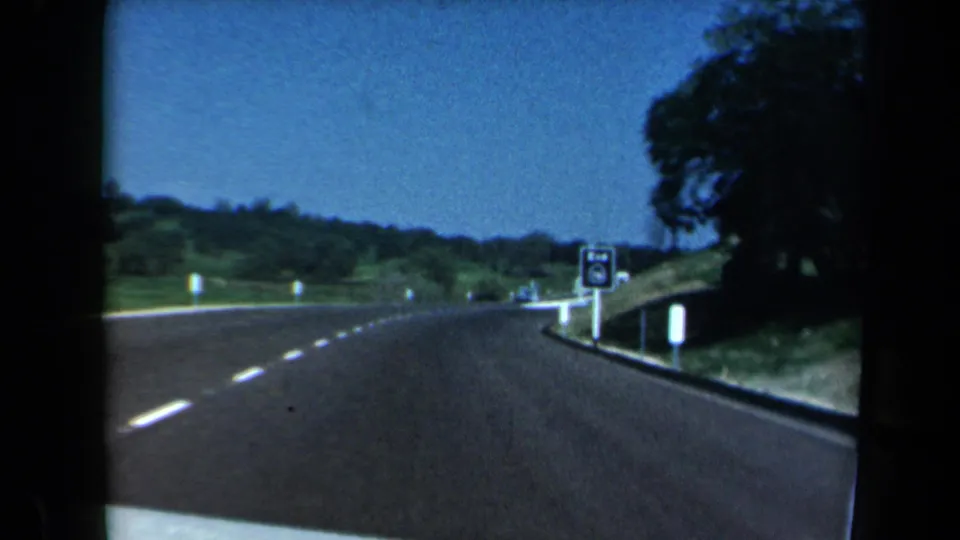 1969 California road footage showing a highway lane, a small route-end sign, green hills, and trees from a moving car.