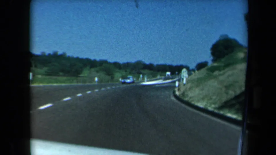 1969 California road-travel frame showing a highway curve from inside a vehicle, with another car ahead and green banks along the shoulder.