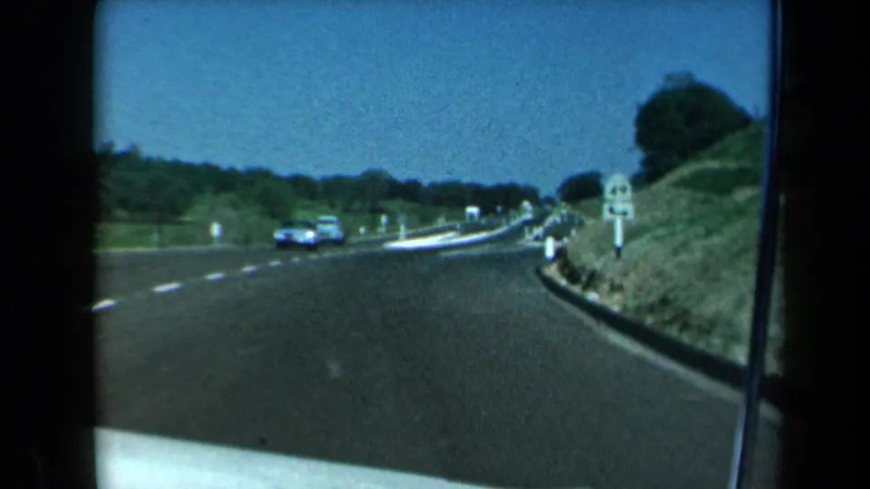 1969 California 8mm highway view from a moving vehicle, with a blue car near the curve and roadside signage on a grassy shoulder.