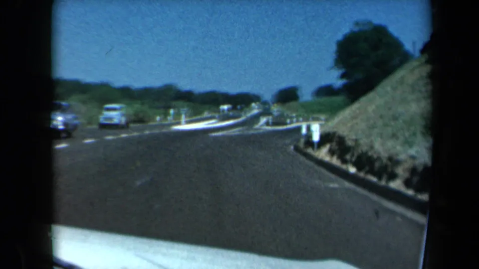 1969 California home-movie frame of a highway bend with cars passing, a roadside sign, and a grassy slope under clear daylight.