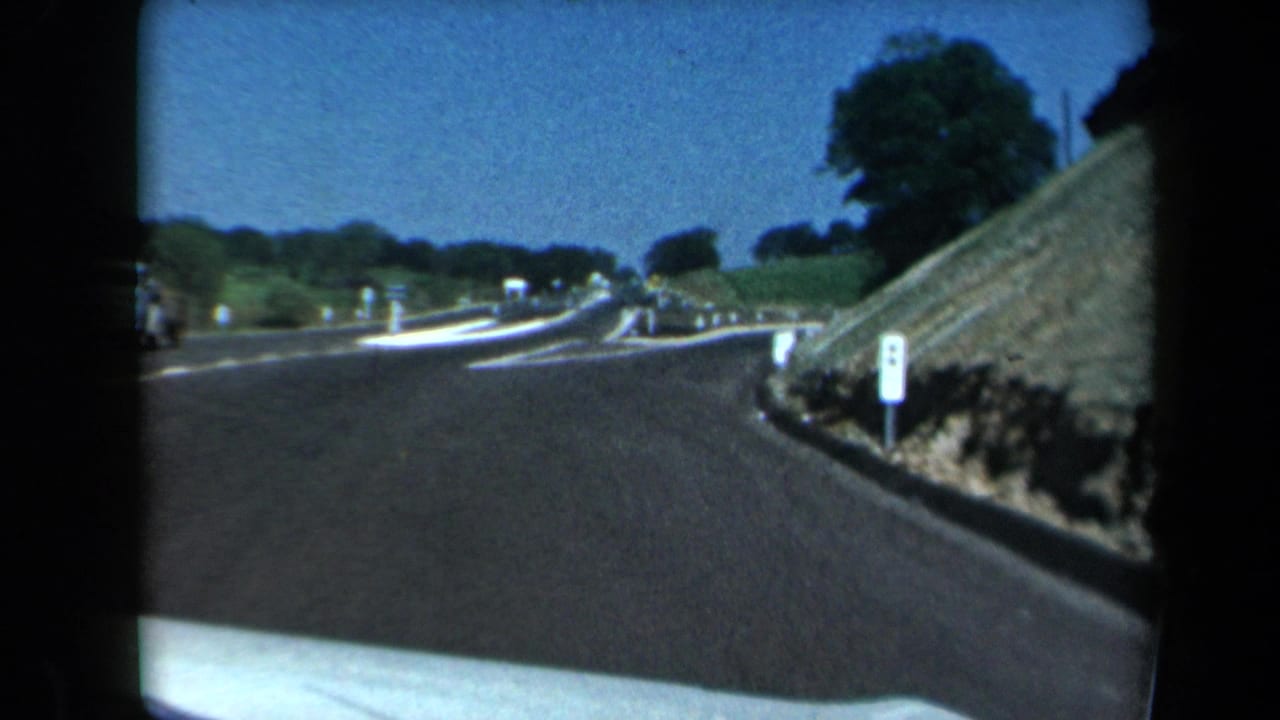 1969 California 8mm view through a windshield as a paved road curves past white roadside posts and a grassy embankment.