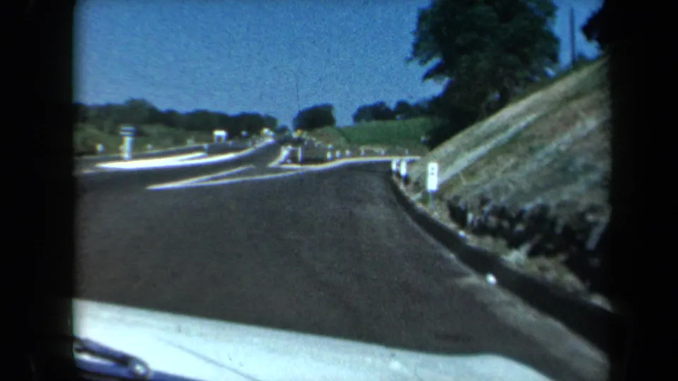 1969 California home-movie frame showing a road bending right with white guide posts, lane markings, trees, and a dashboard edge.