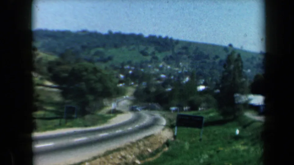 1969 California 8mm frame looking downhill along a winding road toward green hills, trees, and distant structures or vehicles.