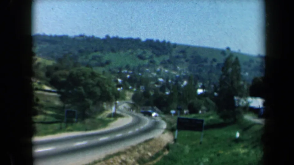 1969 California road-travel frame showing a descending curve, green hillside, roadside signs, and distant vehicles from a car viewpoint.