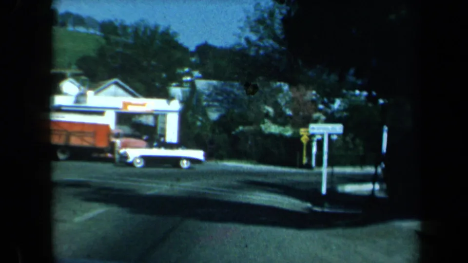 1969 California roadside village view with a white and red car, low building, street signs, trees, and pavement markings.