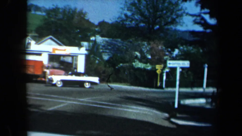 1969 California home-movie frame showing a roadside building, white car with red detail, signs, trees, and a quiet intersection.