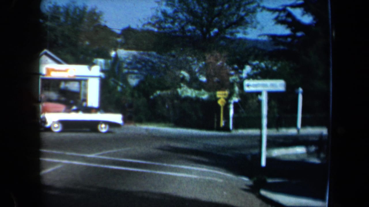 1969 California 8mm frame showing a white and black convertible near a street corner, roadside signs, trees, and a small gas station beyond.