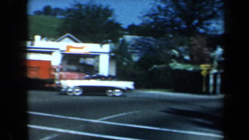 1969 California home-movie frame with a two-tone convertible and red delivery truck moving past a roadside gas station with orange-and-white signage.