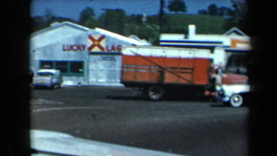 1969 California frame showing a Lucky Lager building, parked compact cars, and a red delivery truck in a roadside commercial area.
