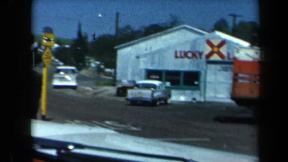 1969 California 8mm frame of a roadside Lucky Lager building with compact cars, a yellow traffic sign, and part of a red truck at frame edge.